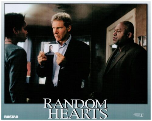 Man in a suit holds a portrait photo, flanked by two other men in a dim hallway; tense moment on a 'Random Hearts' poster.