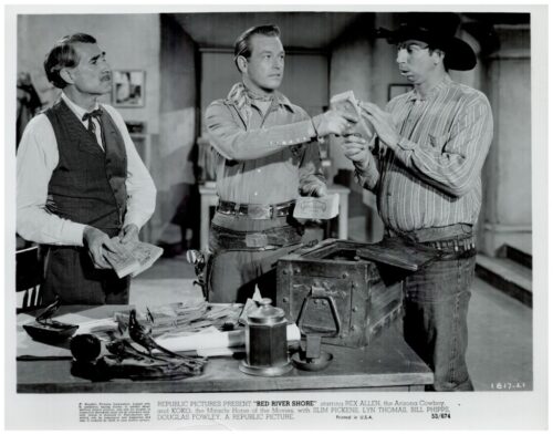 Three men in Western attire count money around a desk with a safe open and scattered cash, a cowboy hat resting on the table.