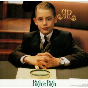 Young boy in a black suit and tie sits at a desk with hands clasped, a magnifying glass on a napkin in front; 'Richie Rich' logo visible.