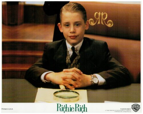 Young boy in a black suit and tie sits at a desk with hands clasped, a magnifying glass on a napkin in front; 'Richie Rich' logo visible.