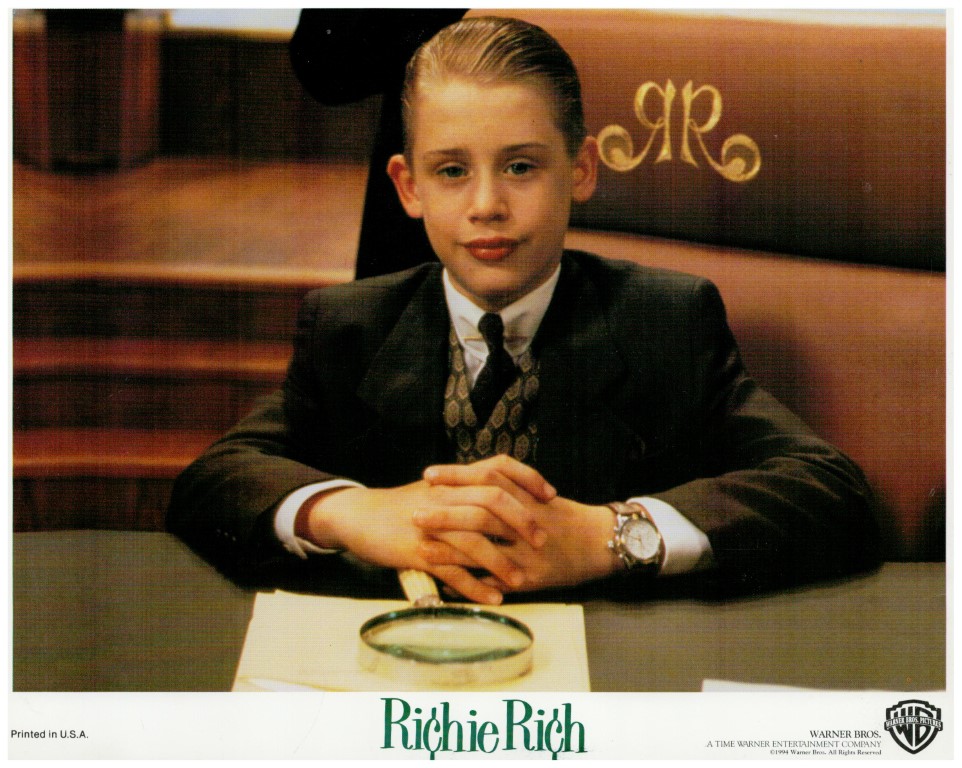 Young boy in a black suit and tie sits at a desk with hands clasped, a magnifying glass on a napkin in front; 'Richie Rich' logo visible.