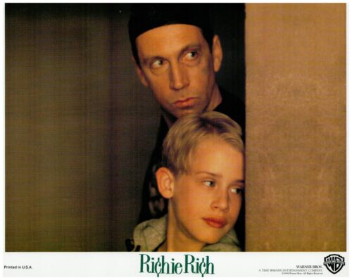 Promotional image from Richie Rich: a blond boy peeks from behind a wall as a man in a black cap looks over from the doorway.
