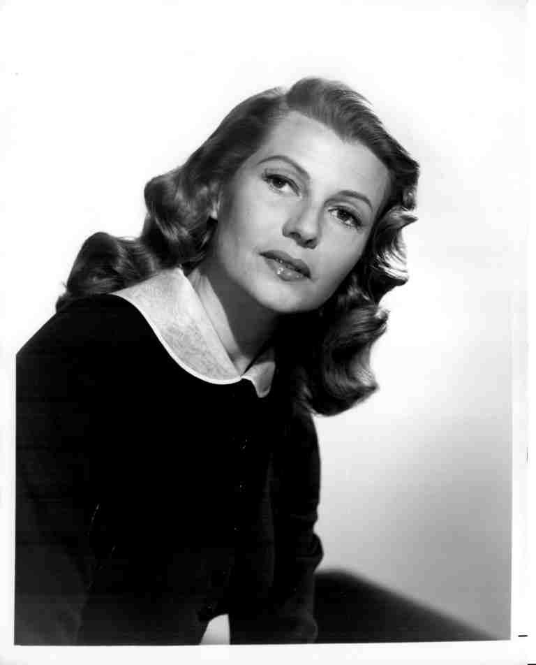 Black-and-white portrait of a woman with wavy hair, wearing a dark outfit and a white collar, looking at the camera.