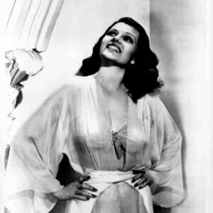 Vintage black-and-white photo of a smiling woman in a satin robe, hands on hips, looking upward by a curtain.