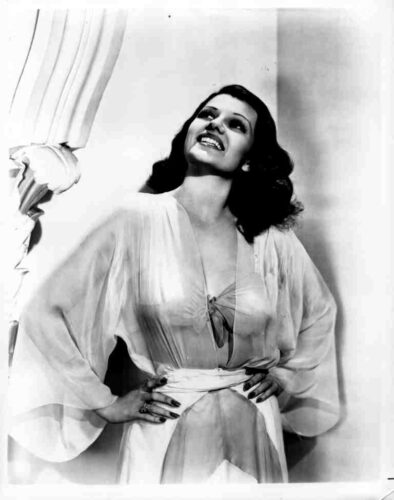 Vintage black-and-white photo of a smiling woman in a satin robe, hands on hips, looking upward by a curtain.