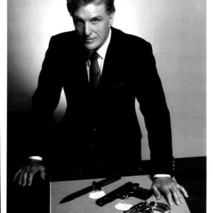 Promotional black-and-white photo of a man in a suit leaning on a desk with guns, from Unsolved Mysteries.