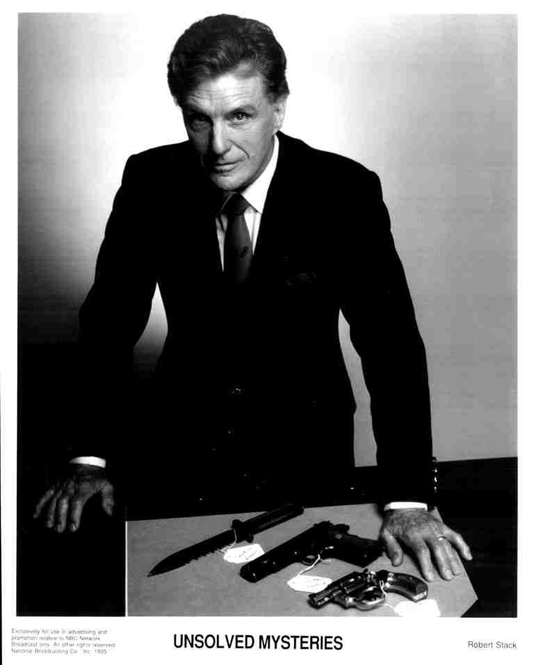 Promotional black-and-white photo of a man in a suit leaning on a desk with guns, from Unsolved Mysteries.