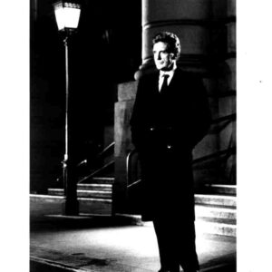 Black-and-white photo of a man in a long dark overcoat and tie standing on a lit sidewalk beside stone steps and a streetlamp at night.