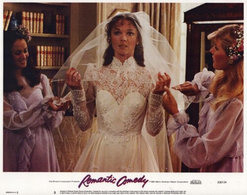 Bride in an ornate lace wedding dress and veil stands center as two lavender-robed bridesmaids adjust her veil.