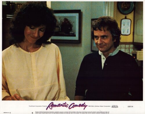 Woman in a pale blouse and a man in a dark sweater smile in a kitchen; a film still labeled 'Romantic Comedy'