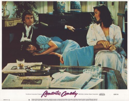Living room scene: a man in a dark suit sits on a couch as a blue-dressed woman lies across his lap, while another woman in a robe sits nearby.