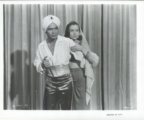 Two performers pose on stage: a man in a turban and white shirt with shiny pants, and a woman in a veil standing close behind him against a striped curtain.