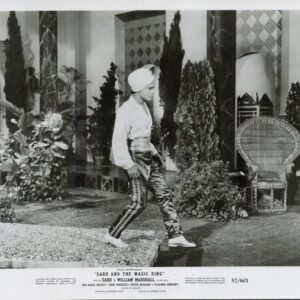 Film still: a man in a turban and white shirt walks across a staged courtyard with plants and a wicker chair in the background.