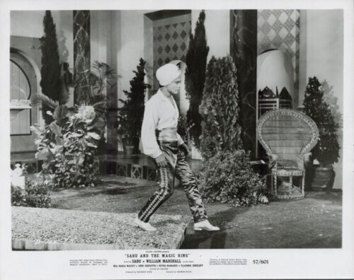 Film still: a man in a turban and white shirt walks across a staged courtyard with plants and a wicker chair in the background.