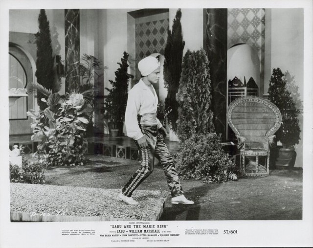 Film still: a man in a turban and white shirt walks across a staged courtyard with plants and a wicker chair in the background.