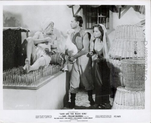 Black-and-white still from a vintage film: a man presents a glowing object to an older seated figure as a woman watches, with woven baskets nearby.