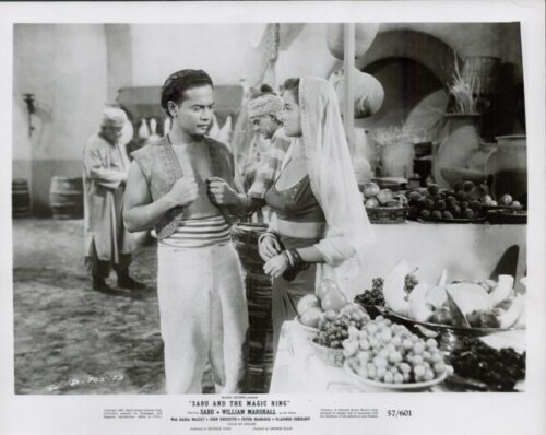 Publicity still from the film 'Sabu and the Magic Ring' showing a man in period dress talking to a veiled woman beside a fruit market stall.