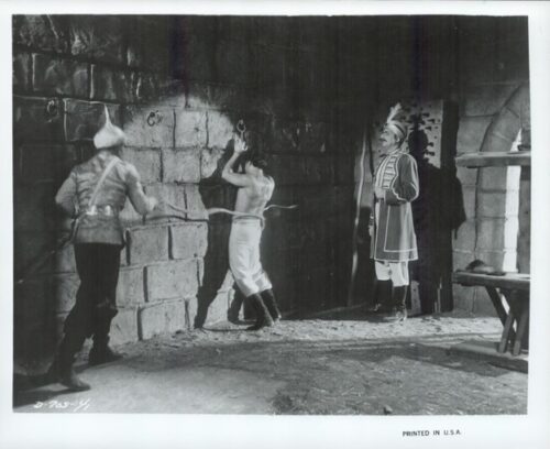 Theatrical scene: two men in period costumes bind a man to a stone wall while a jester-like figure watches.