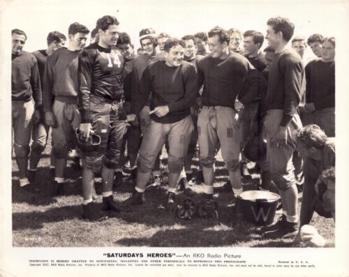 Group of young football players in warmups on a field, smiling at the center; one player wears #14 and holds his helmet.