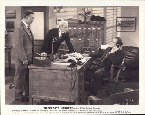Three men in a 1940s office; one sits at a desk on the phone while two others lean in to discuss.