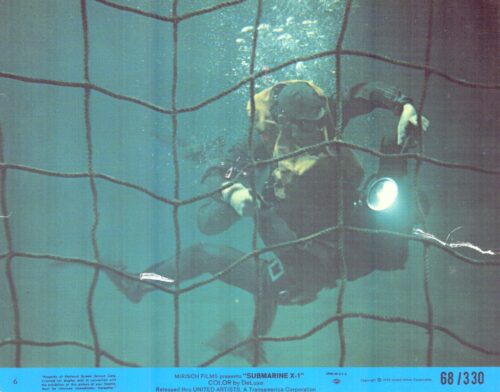 Scuba diver in diving suit and helmet behind a net underwater, flashlight lit in hand. The scene is viewed through a grid-like netting.