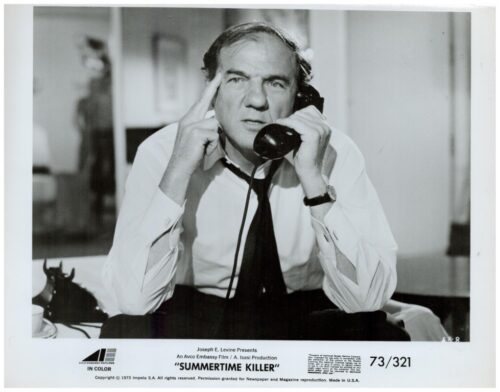 Black-and-white publicity photo of a man in a white shirt and black tie talking on a telephone, resting his finger on his temple, with production text at the bottom.