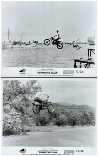 Motorcycle stunt over water: rider mid-air near a dock while a person jumps, boats visible in the harbor background, in black and white.