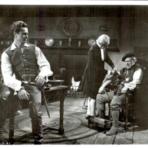 Three men in a Victorian parlor; left man in a vest stands, middle man attends a seated man on the right, with a fireplace in the background.