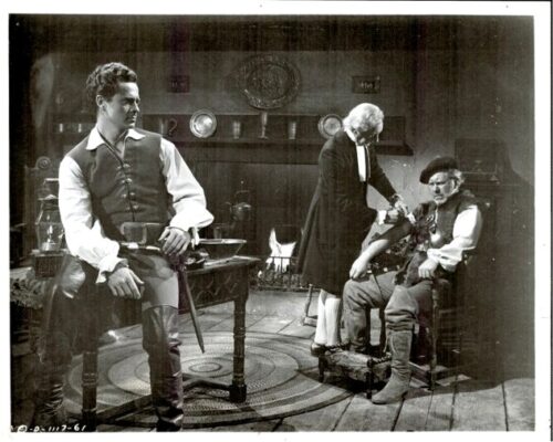 Three men in a Victorian parlor; left man in a vest stands, middle man attends a seated man on the right, with a fireplace in the background.