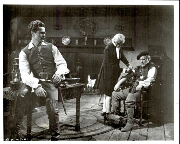 Three men in a Victorian parlor; left man in a vest stands, middle man attends a seated man on the right, with a fireplace in the background.