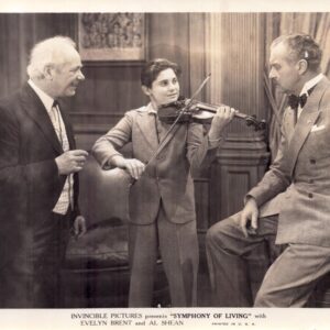 Young boy in a suit plays the violin while two men in suits watch in a wood-paneled room, a vintage studio scene.