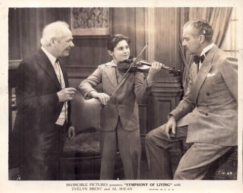 Young boy in a suit plays the violin while two men in suits watch in a wood-paneled room, a vintage studio scene.