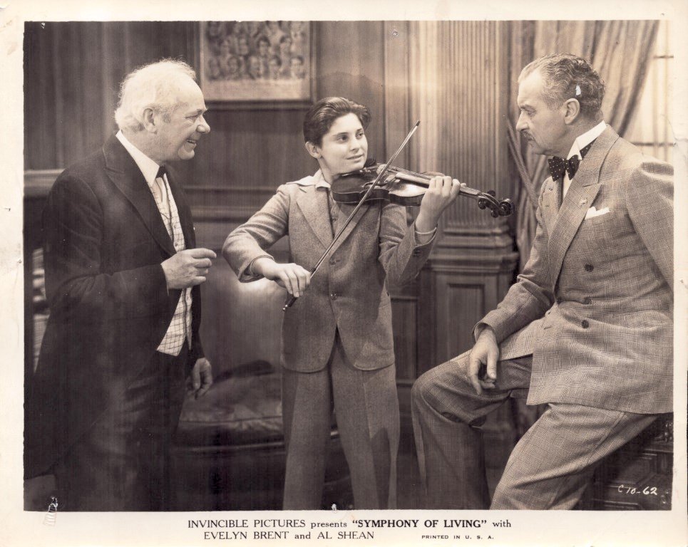 Young boy in a suit plays the violin while two men in suits watch in a wood-paneled room, a vintage studio scene.