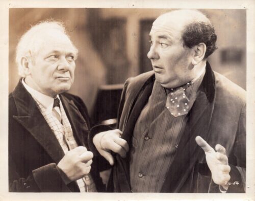 Two older men in vintage attire converse closely, one with white hair and plaid shirt, the other in a striped suit and polka-dot bow tie.