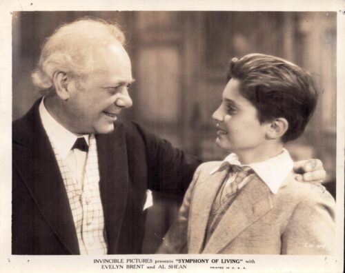 Older man in a suit with his arm around a boy in a light blazer, smiling at each other in a warm moment.