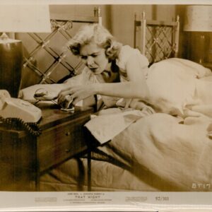 A woman sits on a bed in a bedroom, reaching for a corded telephone on the nightstand beside her.