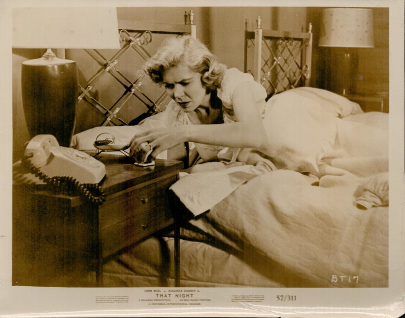 A woman sits on a bed in a bedroom, reaching for a corded telephone on the nightstand beside her.