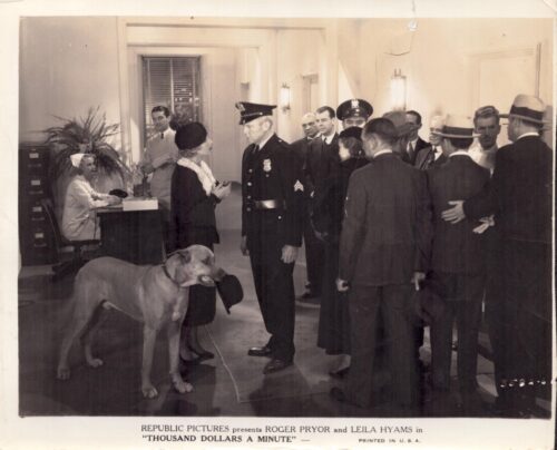 Promotional still for Thousand Dollars a Minute: a uniformed officer faces a couple as a large dog sits nearby in a hotel lobby with a crowd watching.