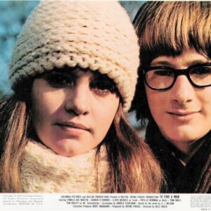 Two young women posing outdoors in winter clothing, one wearing a knit hat and scarf, the other with glasses and a hooded coat.