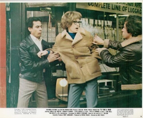 Three people on a city sidewalk; a woman in an oversized tan coat is restrained by two men in leather jackets outside a shop window.