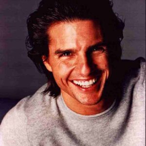 Close-up of a man with dark hair smiling broadly, wearing a light gray crewneck sweater.