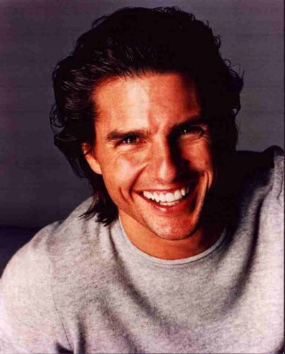 Close-up of a man with dark hair smiling broadly, wearing a light gray crewneck sweater.