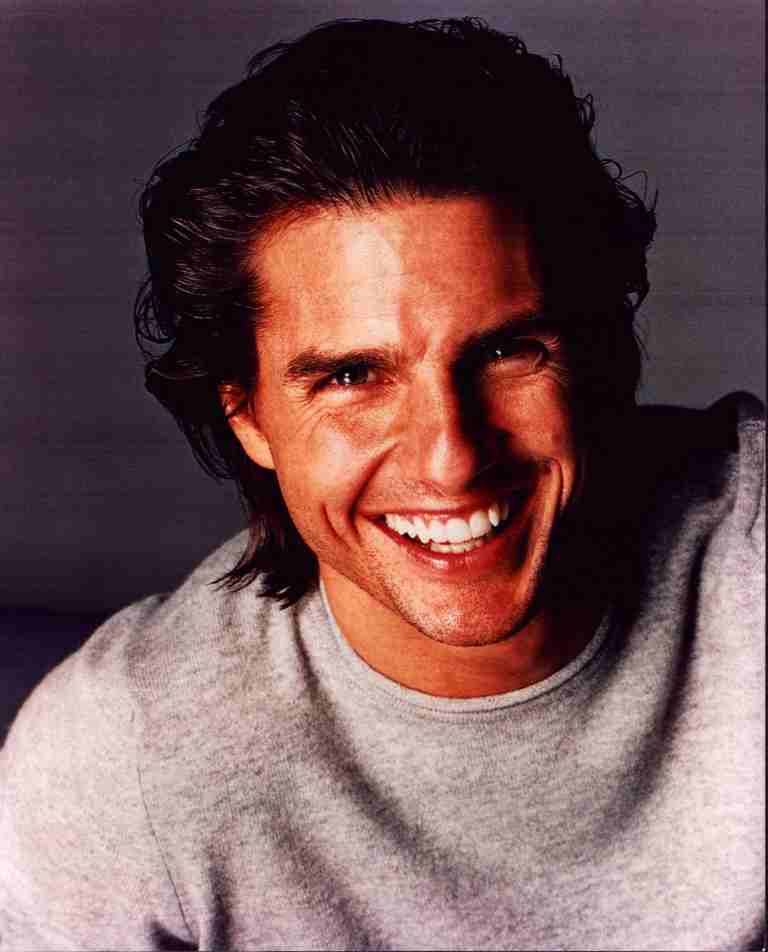 Close-up of a man with dark hair smiling broadly, wearing a light gray crewneck sweater.
