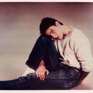 Young man sits on the floor with his head resting on his knee, wearing a light sweater and jeans.