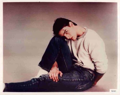 Young man sits on the floor with his head resting on his knee, wearing a light sweater and jeans.