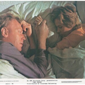 A man and a woman lie in bed facing each other, partially covered by blankets, in a tense scene.