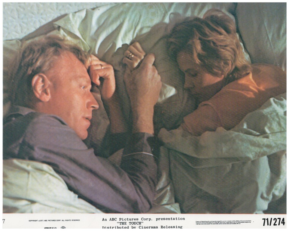 A man and a woman lie in bed facing each other, partially covered by blankets, in a tense scene.