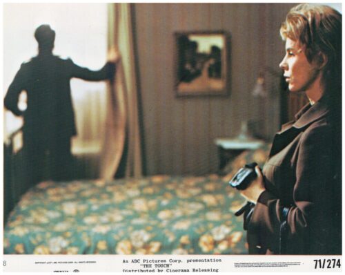 Film still: a woman in a brown coat holds a camera, facing right, as a shadowy man pulls back a curtain in the background.