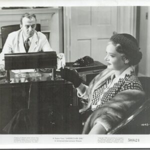 Black-and-white still of a man in a white coat at a desk, facing a woman in a checked coat and fur stole seated nearby, in a vintage setting.