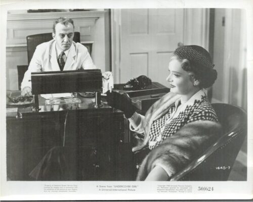 Black-and-white still of a man in a white coat at a desk, facing a woman in a checked coat and fur stole seated nearby, in a vintage setting.